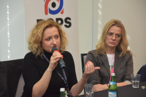 dwie kobiety siedzą przy stole, jedna z nich trzyma mikrofon