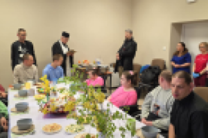 ludzie siedzą przy stole, duchowny stoi i czyta