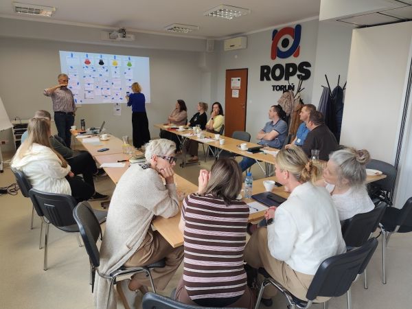 Uczestnicy szkolenia siedzą przy stołach ustawionych w podkowę w sali konferencyjnej ROPS w Toruniu. Prowadzący stoi przy...
