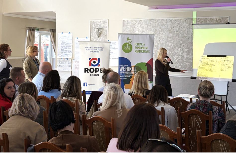 Sala konferencyjna podczas spotkania. Kobieta przemawia do siedzącej publiczności, trzymając mikrofon i wskazując planszę...