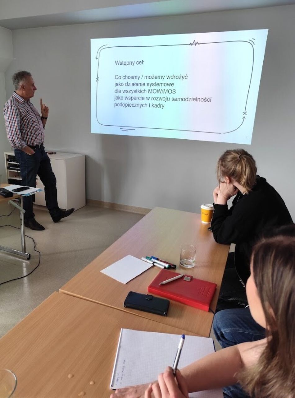 Mężczyzna, Prezes Fundacji Samodzielni Robinsonowie, podczas warsztatów w sali szkoleniowej wskazuje na prezentację...